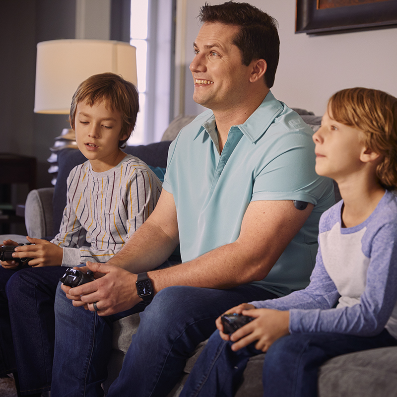 Mann spiller videospill med barna mens han har en implanterbar CGM på armen.